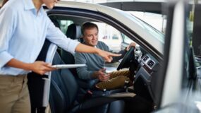 A man sitting in a car being instructed on how to use the vehicle by a woman with a tablet