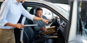 A man sitting in a car being instructed on how to use the vehicle by a woman with a tablet