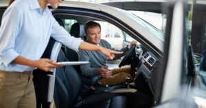 A man sitting in a car being instructed on how to use the vehicle by a woman with a tablet