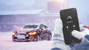 A person's hands pressing the start button for a remote start on their phone for their car which is covered in snow on a cold day